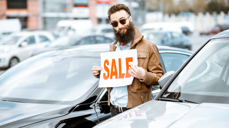 A man wearing sunglasses and a brown jacket is standing in the middle of a bunch of cars holding a sign that says "sale".