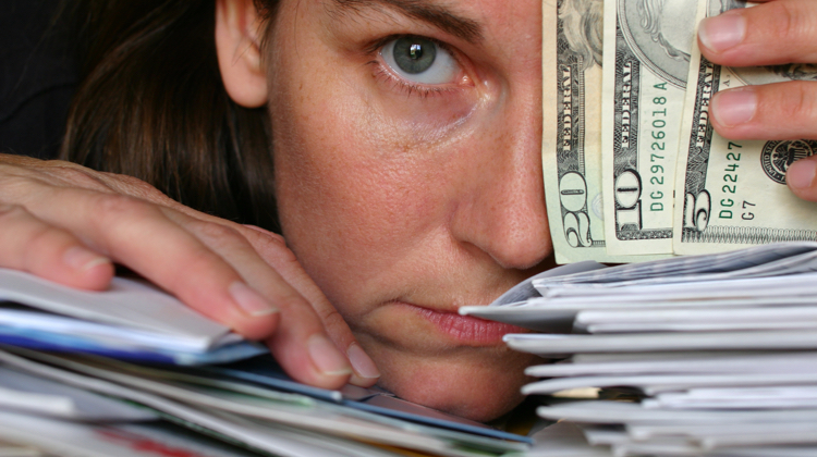 Face rests on a stack of mail with one eyeball covered by a hand holding one five, one ten, and one twenty dollar US bills.