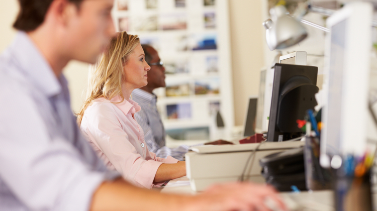 Three workers sit side-by-side working on the computer; the man in the foreground is blurred and focus is on blonde woman in pink shirt.