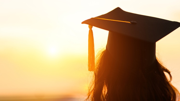Silhouette of a woman wearing a graduation cap staring into a setting sun; portfolio school isn't the only way to become a copywriter.