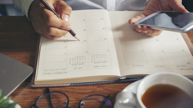 Looking at a pair of hands, the left hand holding an iPhone, the right holding a pencil and writing in a planner with a cup of coffee and glasses in front of the planner.