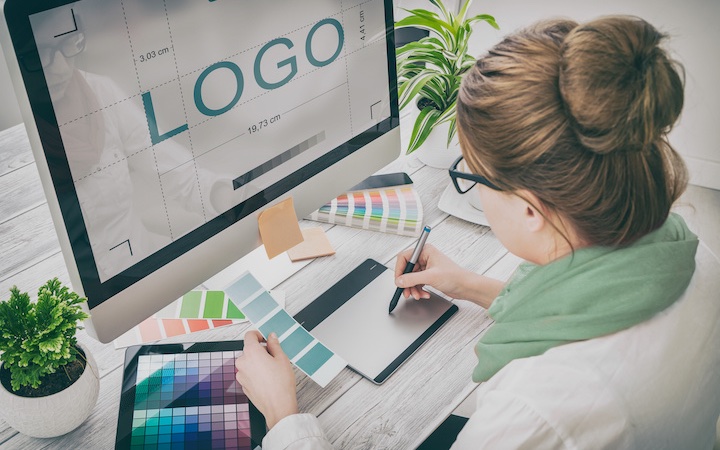 A woman with hair in bun, glasses, and scarf uses a tablet and a stylus to design a logo while the word "logo" appears on the monitor in front of her.
