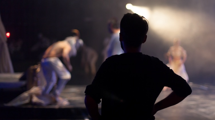 Silhouette of a man on stage with hands on his hips looking at performers in the spotlight.