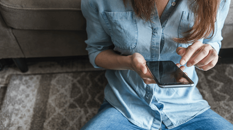 Image is cropped showing a person's torso wearing a denim top and jeans as they sit on a carpet holding a phone and pointing a finger at the screen.