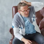 man wearing glasses and a cardigan and sitting in a leather chair is scratching his forehead and looking bewildered