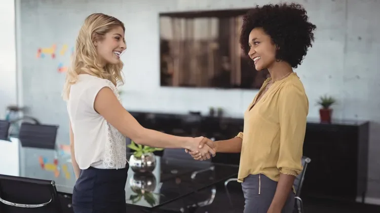 Two women in their thirties shaking hands and smiling in a modern conference room