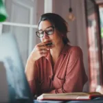 Woman wearing glasses and a pink button-down shirt sits before her laptop looking contemplative with her chin in her hands