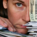 Face rests on a stack of mail with one eyeball covered by a hand holding one five, one ten, and one twenty dollar US bills.