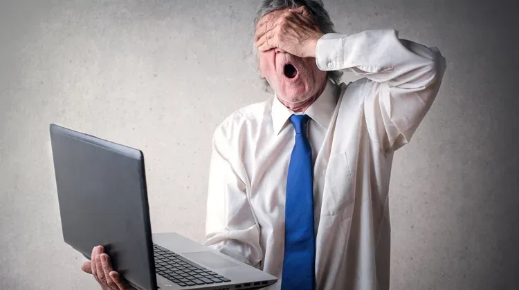 Older man in white dress shirt and blue tie sis in front of a computer with his hand covering his face and his mouth open in surprise.