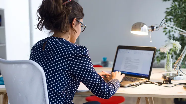 Looking over the shoulder of a woman in a navy polka dress, with glasses on, works on her laptop computer.