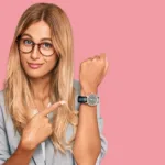Woman in blazer stands against a salmon background pointing at her watch