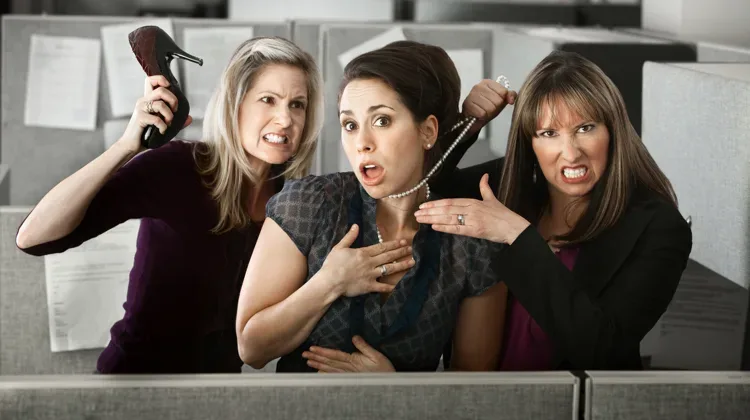 Three women are all dressed in black outfits as one on the left threateningly holds a heel up to the one in the middle while the woman on the right pulls at the middle woman's pearl necklace.