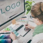 A woman with hair in bun, glasses, and scarf uses a tablet and a stylus to design a logo while the word "logo" appears on the monitor in front of her.
