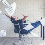 Man, who is sitting in front of a light-filled window, leans back in a chair with his arms outstretched as papers scatter all around from his hands