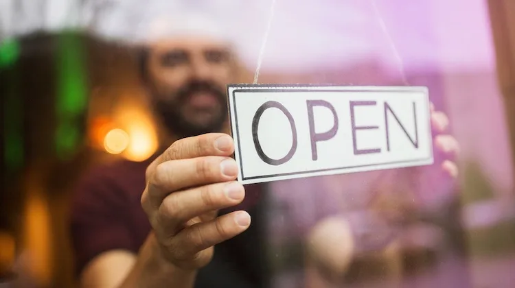 An out-of-focus bearded man flips a paper sign to "open" in the window of a small business.